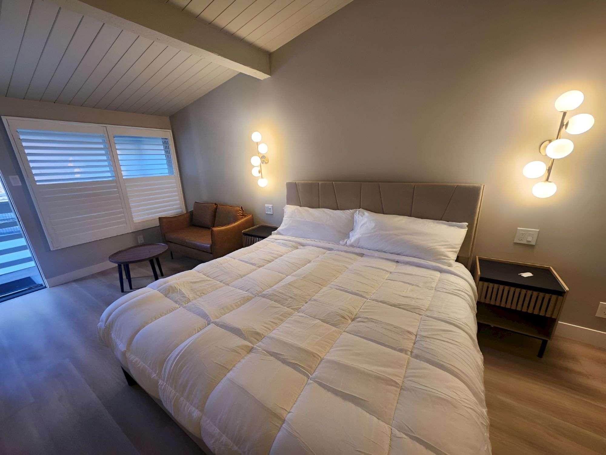 A modern, comfy bedroom with a large bed, soft white comforter, nightstands, warm lighting, and a small seating area by the window.