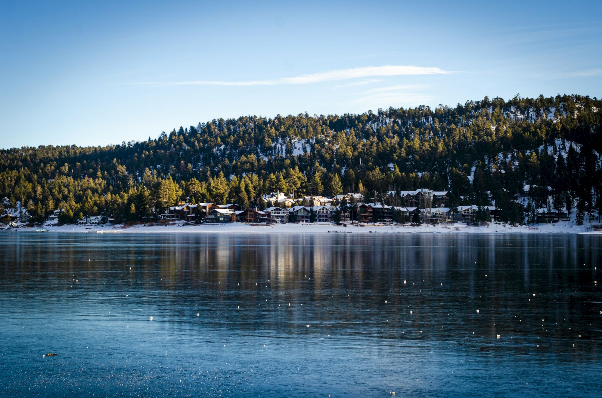 Snowy hillside village along a calm lake, reflections ripple on the water, evergreen trees, clear blue sky, serene winter scene.
