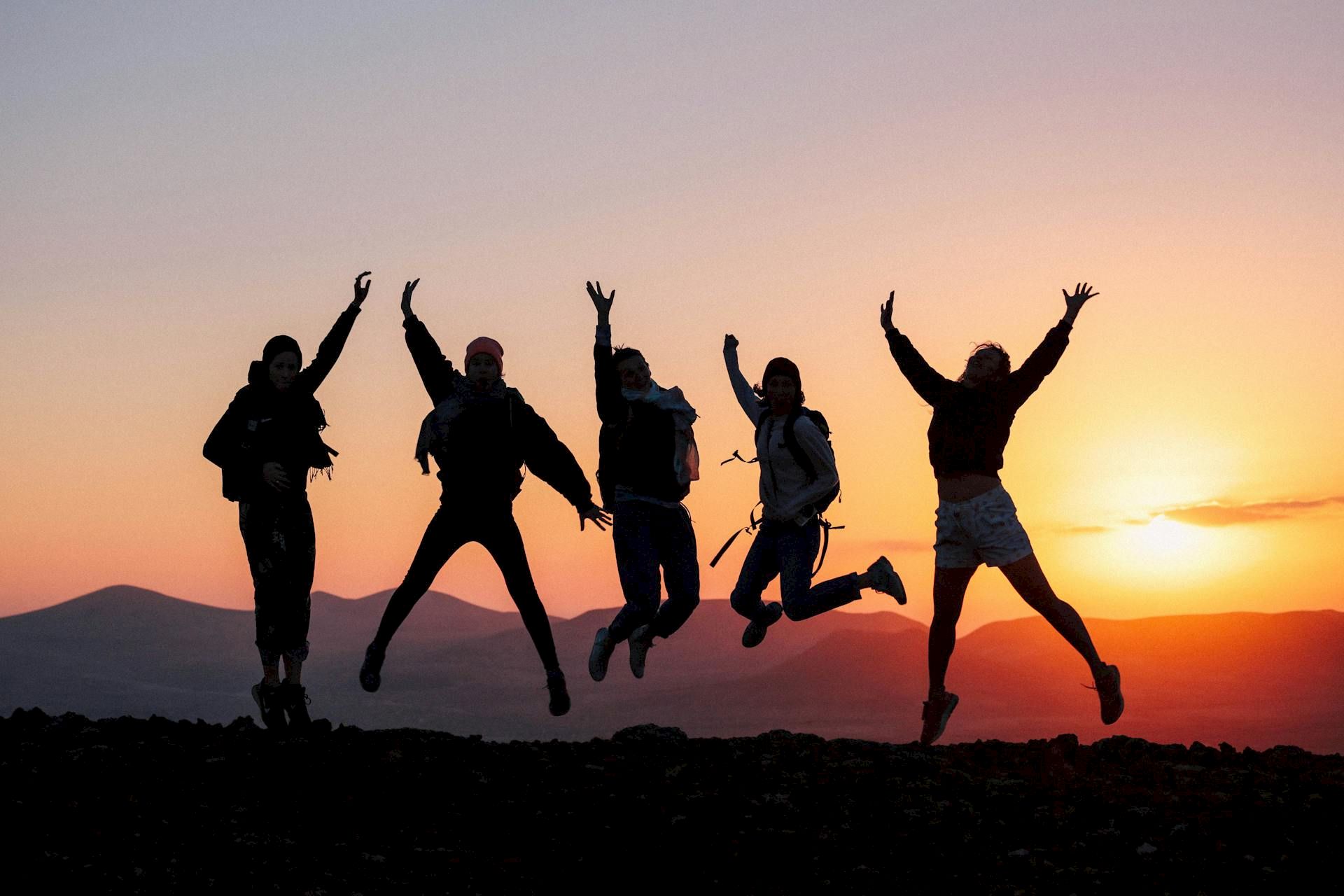 Silhouetted hikers jump for joy on a mountain at sunset, celebrating together against a glowing sky.
