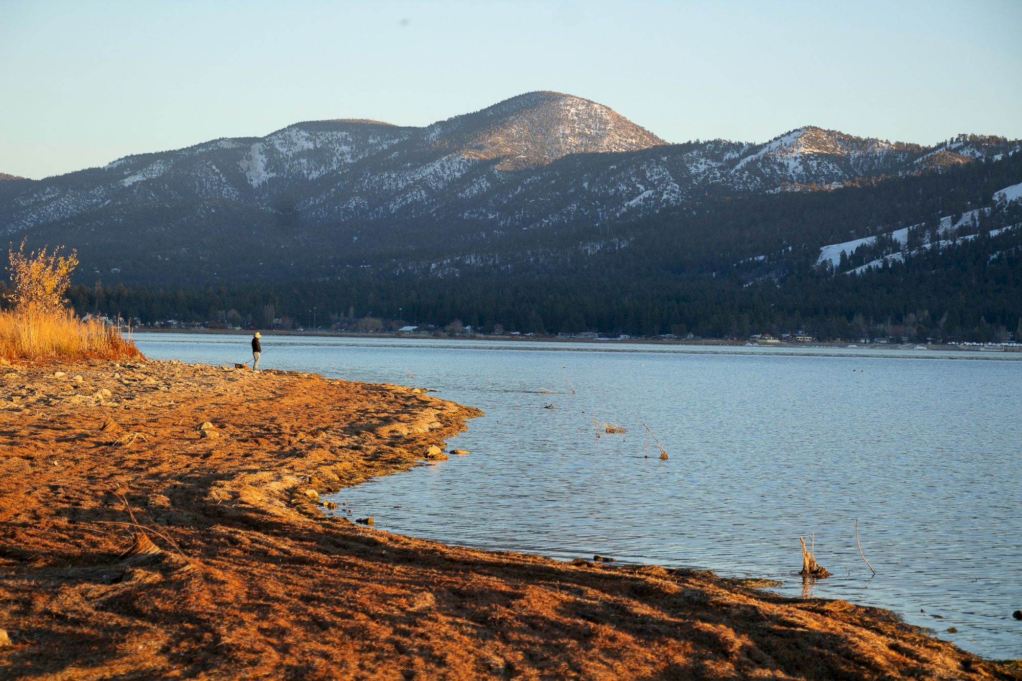 A lakeside scene with calm water, a sandy shore, distant mountains, and a person walking along the edge at sunset.