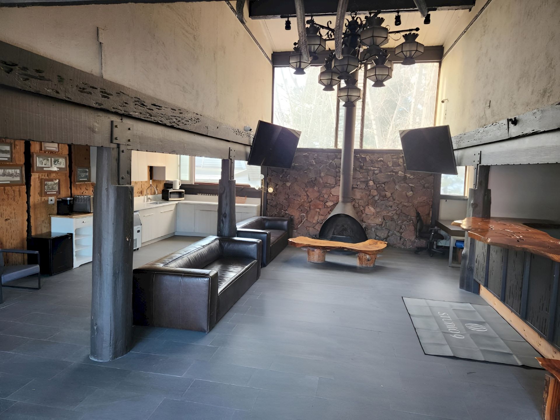 Industrial-style lounge kitchen with two black couches, a stone fireplace, pendant lights, and large windows.