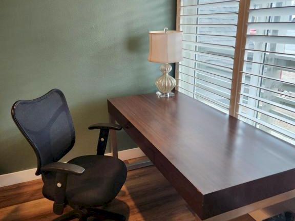 A small home office setup with a dark wooden desk, a black ergonomic chair, a green wall, and a window with white blinds letting in light.