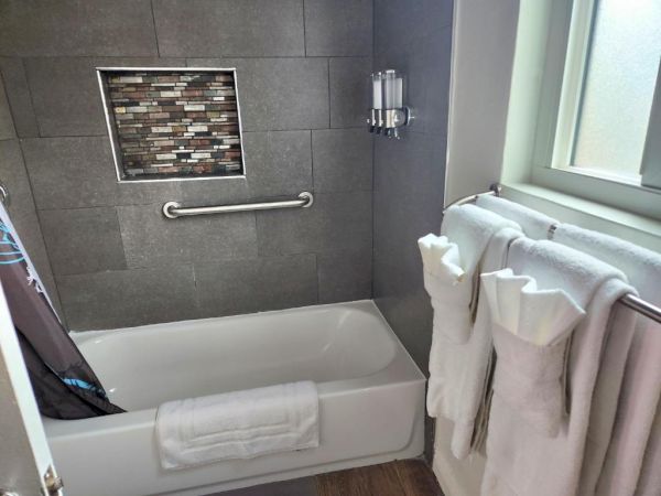 A modern bathroom with a white bathtub, gray tiled walls, a recessed shelf, a grab bar, and fluffy white towels by a window.