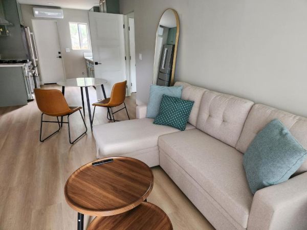 A modern living space with a light sofa, mint cushions, round wooden coffee tables, a small dining area, and a mirror in a bright, open room.