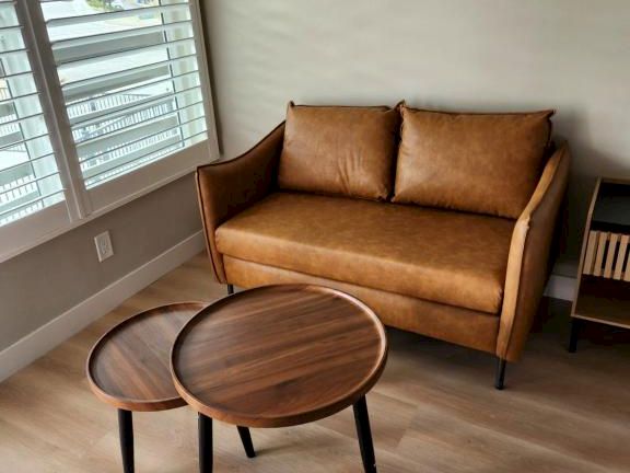 A cozy living area with a tan mid-century sofa, two round nesting coffee tables, light wood floors, and a large window with blinds.