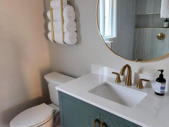 A modern bathroom with a teal vanity, white countertop, round mirror, gold fixtures, a wall-mounted towel rack, and a white toilet.