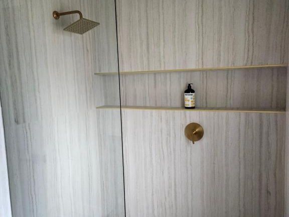 A modern glass-front shower with a wall-mounted rain shower, a handheld bar, and built-in shelves with a small bottle, light gray tile, and a light design.