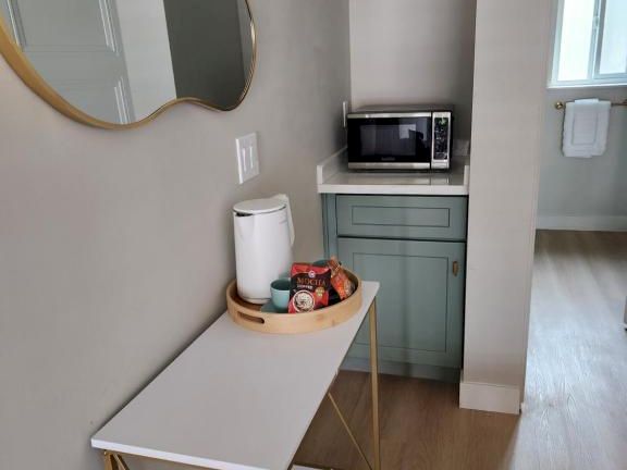 A small entry nook with a narrow console table, a round mirror, a top shelf with a microwave, a kettle, a basket of fruit, and pastel cabinet under, in a tidy space.