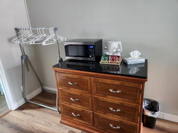 A small kitchenette with a microwave, tissue box, and toaster on a black countertop atop a wooden dresser, with a metal drying rack and trash can nearby.