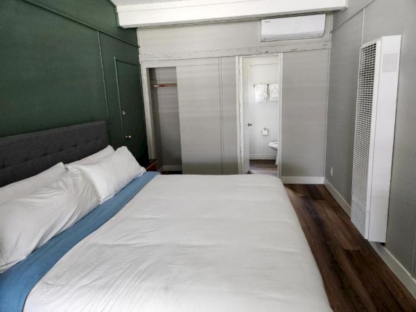 A modern hotel room with a large bed, gray walls, a built-in headboard, and a frosted glass closet door, plus a small bathroom visible in the back.
