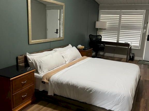 A cozy hotel-style bedroom with a queen bed, two wooden nightstands, a large mirror, and a seating area by the window, under a sloped ceiling.