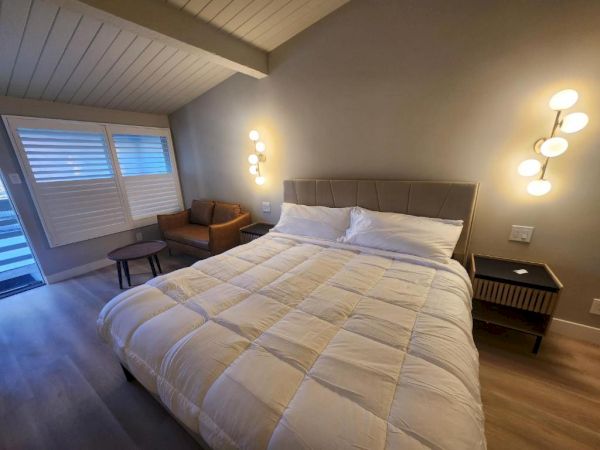 A cozy bedroom with a large bed, white duvet, two matching nightstands, a small seating area by the window, and warm wall lights.