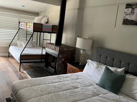 A cozy, modern bedroom with a queen bed in the foreground, a wood-burning stove and bunk area in the back, and soft natural light.