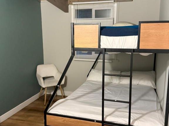 A small bedroom with a metal bunk bed, white mattresses, a wooden twin above, a wooden chair, green wall on left, and a wooden floor under a low ceiling.
