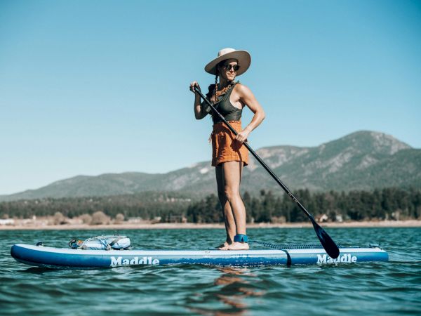 A person stands on a blue paddleboard, wearing a hat and life vest, holding a long paddle, with mountains in the background over calm water.