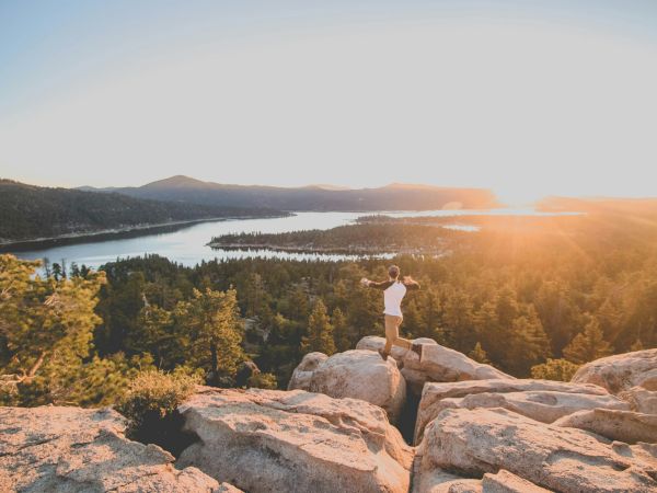 A person stands on rocky cliffs overlooking a lake and forest at sunset, capturing the moment as the sun spills orange light across the scene.