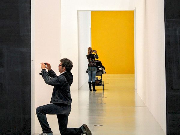 A person kneels on one knee, taking a photo of another person seated in a yellow-walled doorway framed by white walls.