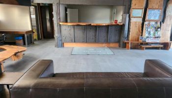 A rustic lounge area with brown leather seating, wooden walls, a bar counter, benches, and a doorway in a cabin-like interior.