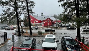 Cars parked in a flooded lot by a red building and trees; rain slicks the ground, creating reflections and a gray sky.
