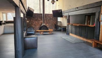 A modern loft-style living space with dark wooden beams, a central brick fireplace, black leather sofas, a round coffee table, and a bar area.
