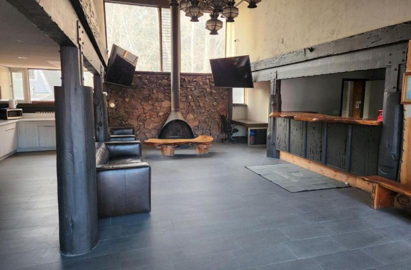 A modern loft-style living space with dark wooden beams, a central brick fireplace, black leather sofas, a round coffee table, and a bar area.