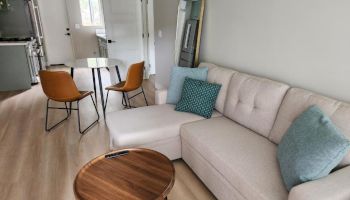 A cozy living area with a beige sectional, teal pillows, a round wood coffee table, a white wall, a tall mirror, and a small dining area with two orange chairs.