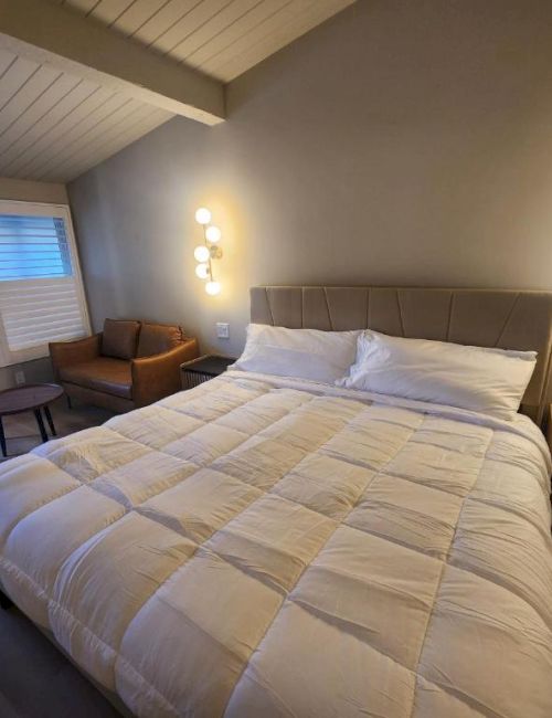 A modern hotel room with a large bed, neutral tones, two nightstands, a cozy chair and table, and warm wall lighting; bright windows on the left.