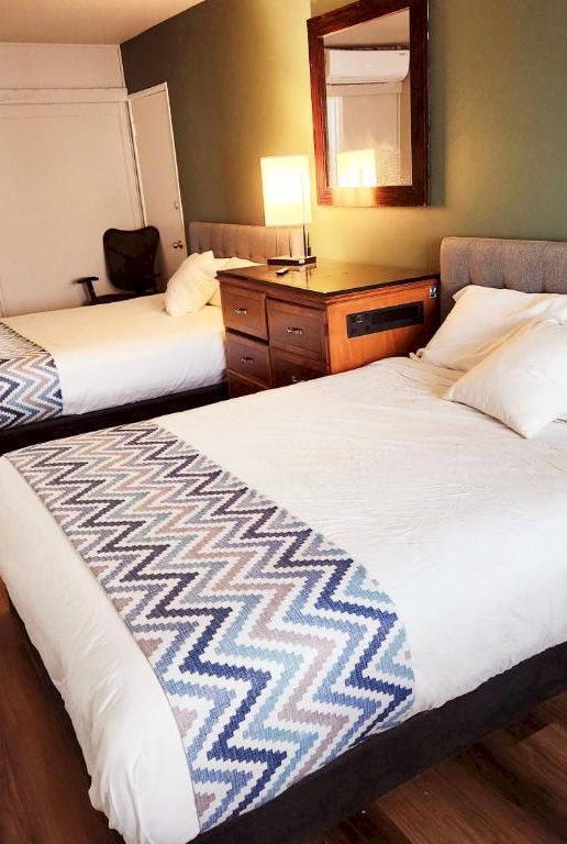 A modern hotel room with two queens, stylish zigzag runners, wooden floors, green accent wall, and bedside lamps beside a padded headboard.