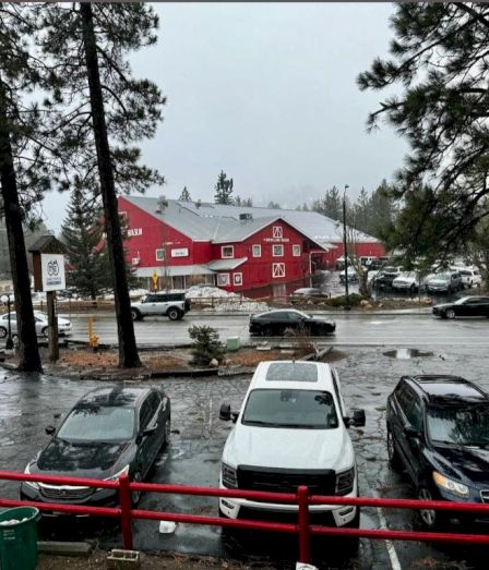 People drive through a parking lot in heavy rain with red wooden buildings in the background, trees framing the scene.