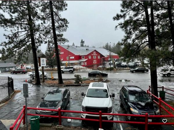 People drive through a parking lot in heavy rain with red wooden buildings in the background, trees framing the scene.
