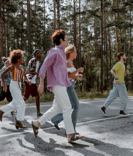 A diverse group of friends cross a sunny forest road together, chatting and walking briskly.