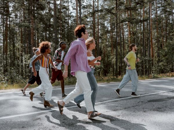 A diverse group of friends cross a sunny forest road together, chatting and walking briskly.