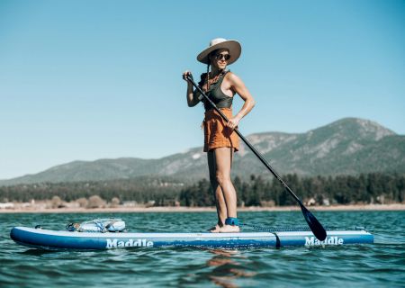 A person standing on a paddleboard or surfboard in the water, with mountains in the background and a clear blue sky.