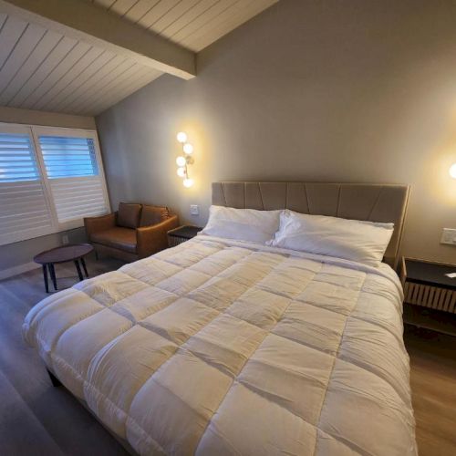 A modern bedroom with a large bed, white comforter, twin nightstands, a brown leather chair by the window, and wall-mounted lamps.