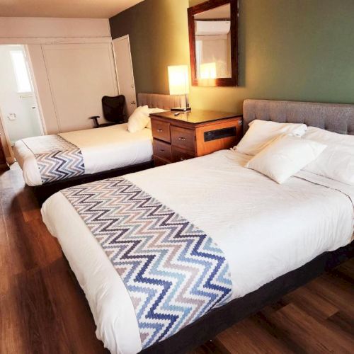 A hotel room with two double beds, chevron runners, wooden floor, green accent wall, nightstands, lamp, and a wall mirror.