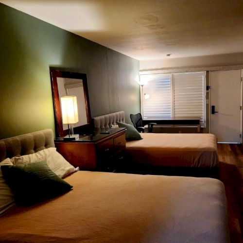 A dim hotel room with two large beds, a green accent wall, a nightstand lamp, and a window at the far end, cozy but sparsely furnished.