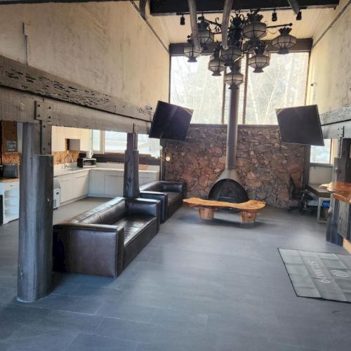 A spacious lounge/bar area with black leather couches, a round wooden table, mounted TVs, a brick wall, and a rustic wooden bar on the right.