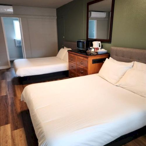 Two neatly made beds in a small hotel room, with a wooden dresser, mirror, mini fridge area, and a windowed door at the back.
