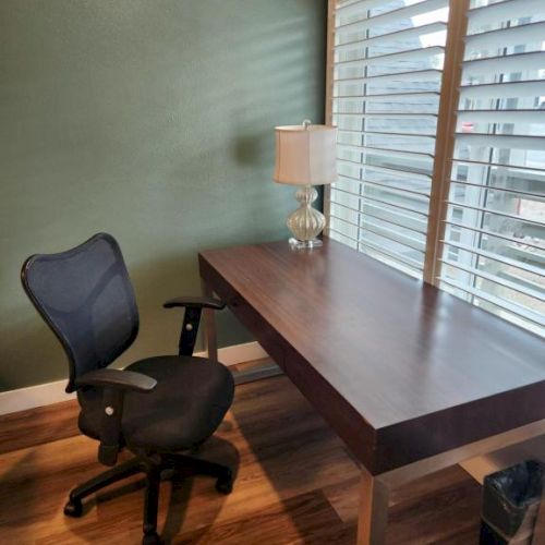 A simple home office with a dark wood desk by a window, a black ergonomic chair, a glass-topped lamp, and green wall color.