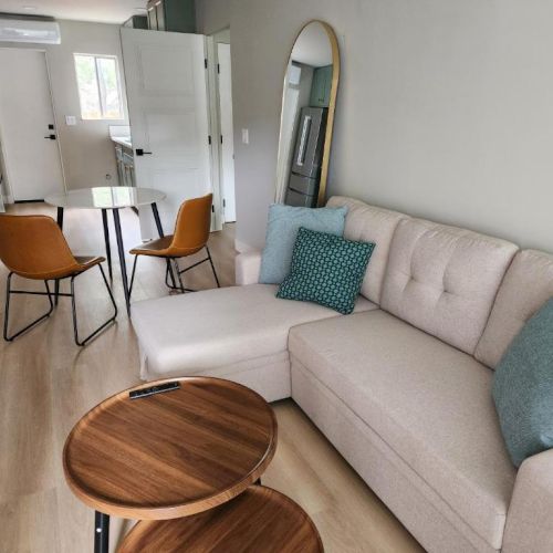 A modern, airy living space with a beige sectional sofa, teal pillows, round wooden coffee tables, a small dining area, and a mirror by the wall.