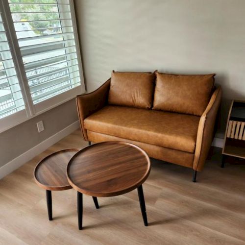 A cozy living room with a tan leather loveseat, two round wooden coffee tables, large windows with blinds, and light wood flooring.